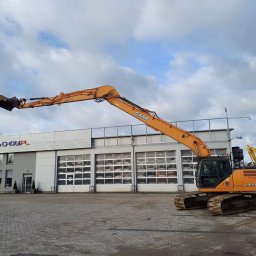 CHOGI SPÓŁKA Z OGRANICZONĄ ODPOWIEDZIALNOŚCIĄ - Pomarańczowa koparka CASE CX210D na tle szarego budynku z logo CHOGI.PL i bramami garażowymi, obok zaparkowane inne maszyny budowlane.