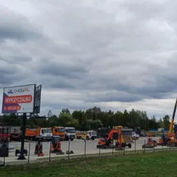 Plac wypożyczalni sprzętu budowlanego Chogi.pl z widocznymi koparkami, podnośnikami koszowymi i samochodami ciężarowymi na tle pochmurnego nieba.