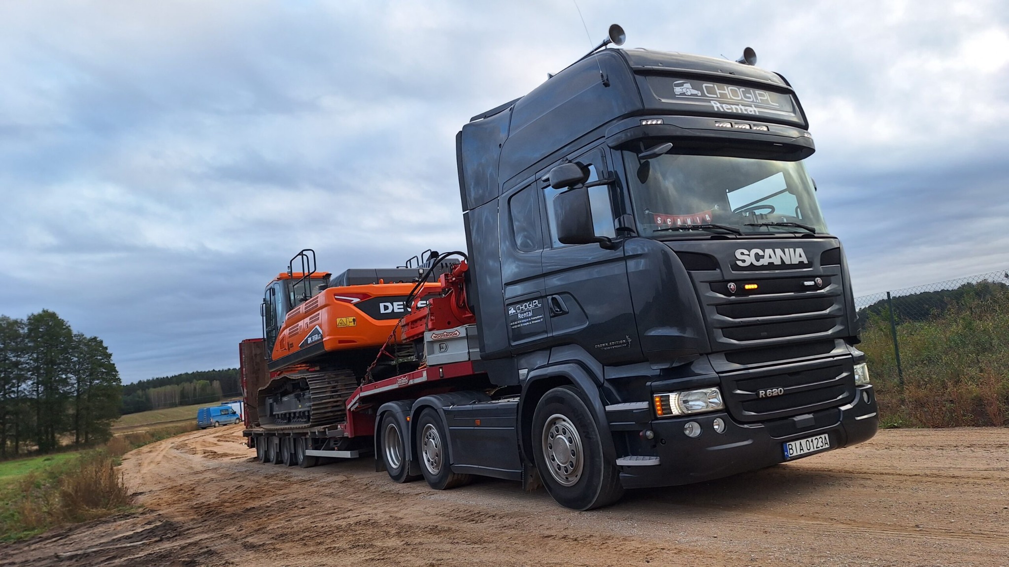 Czarny ciągnik siodłowy Scania R620 przewozi na niskopodwoziowej platformie pomarańczową koparkę gąsienicową Doosan DX 255LC po piaszczystej drodze polnej, widoczne logo 'CHOGIPL Rental' na kabinie.