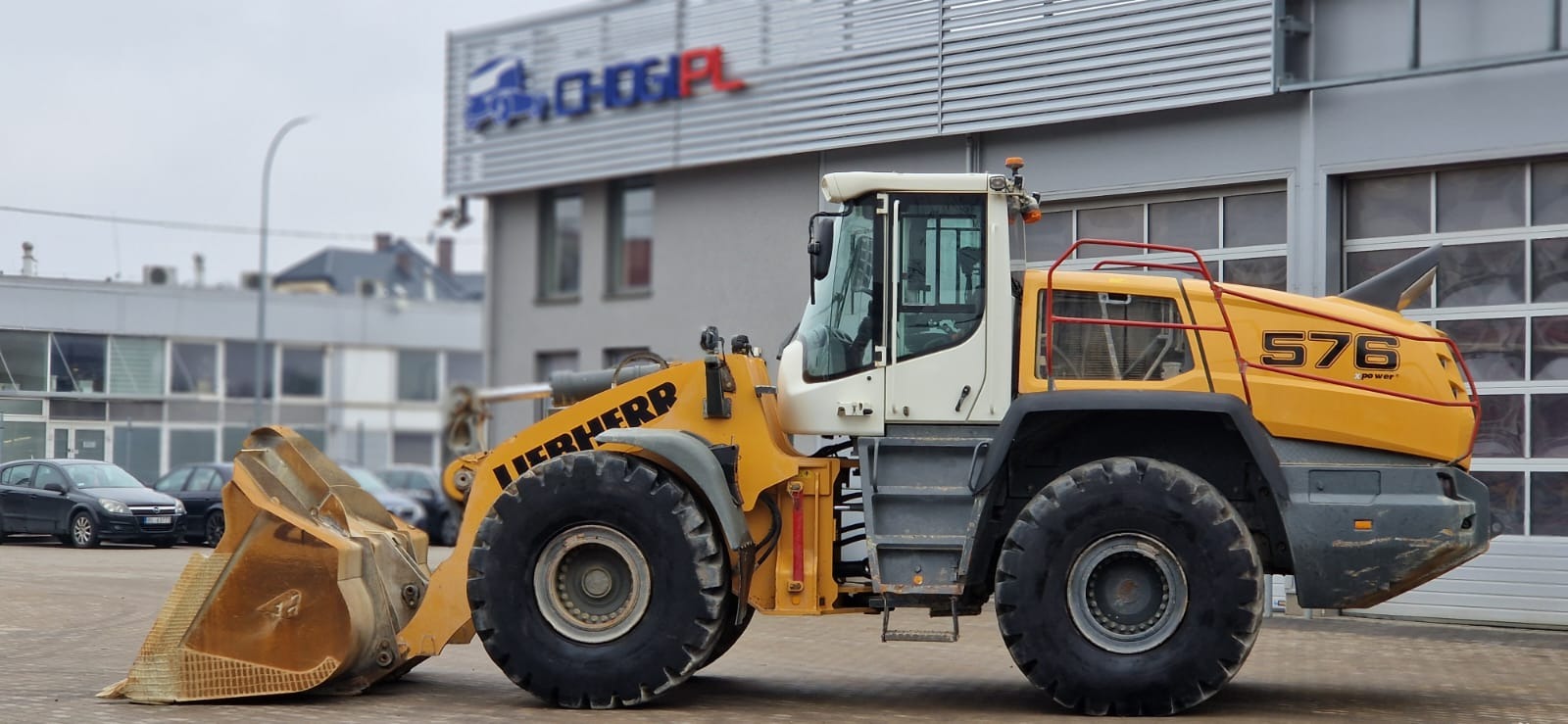 Żółta ładowarka kołowa Liebherr 576 z łyżką, stojąca na placu przed budynkiem z logo firmy CHOGI.PL, na tle szare niebo.