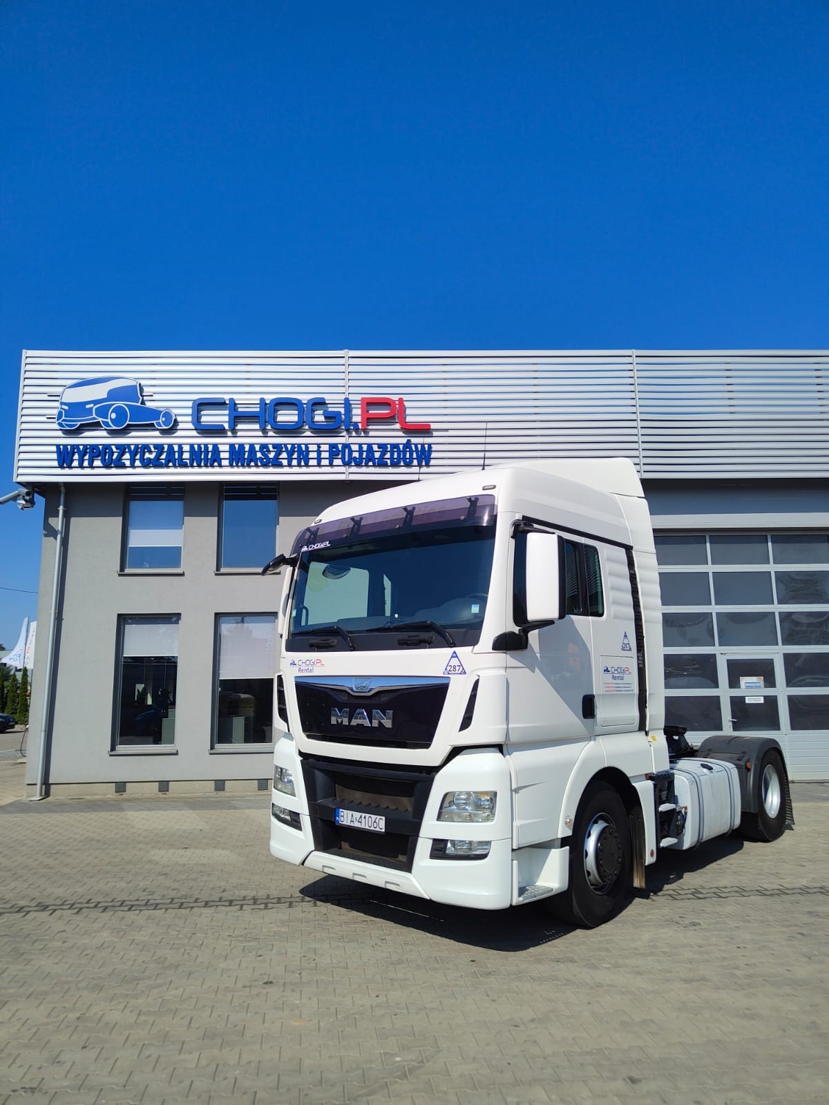 Biała ciężarówka MAN przed budynkiem wypożyczalni maszyn i pojazdów z logo firmy CHOGI.PL na tle błękitnego nieba.