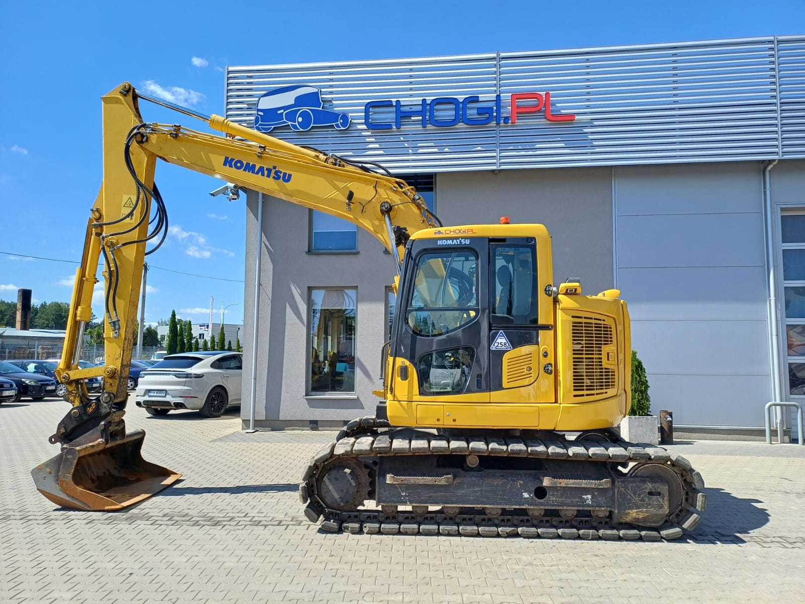 Żółta koparka Komatsu z łyżką na tle budynku z logo firmy, widok z boku, na pierwszym planie kostka brukowa, w tle zaparkowane samochody.