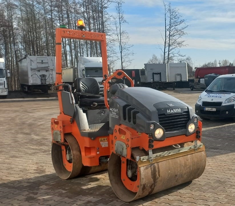 Pomarańczowo-szary walec drogowy HAMM na placu, z włączonymi światłami i żółtym ostrzegawczym światłem na górze, w tle zaparkowane ciężarówki i furgonetka serwisowa.