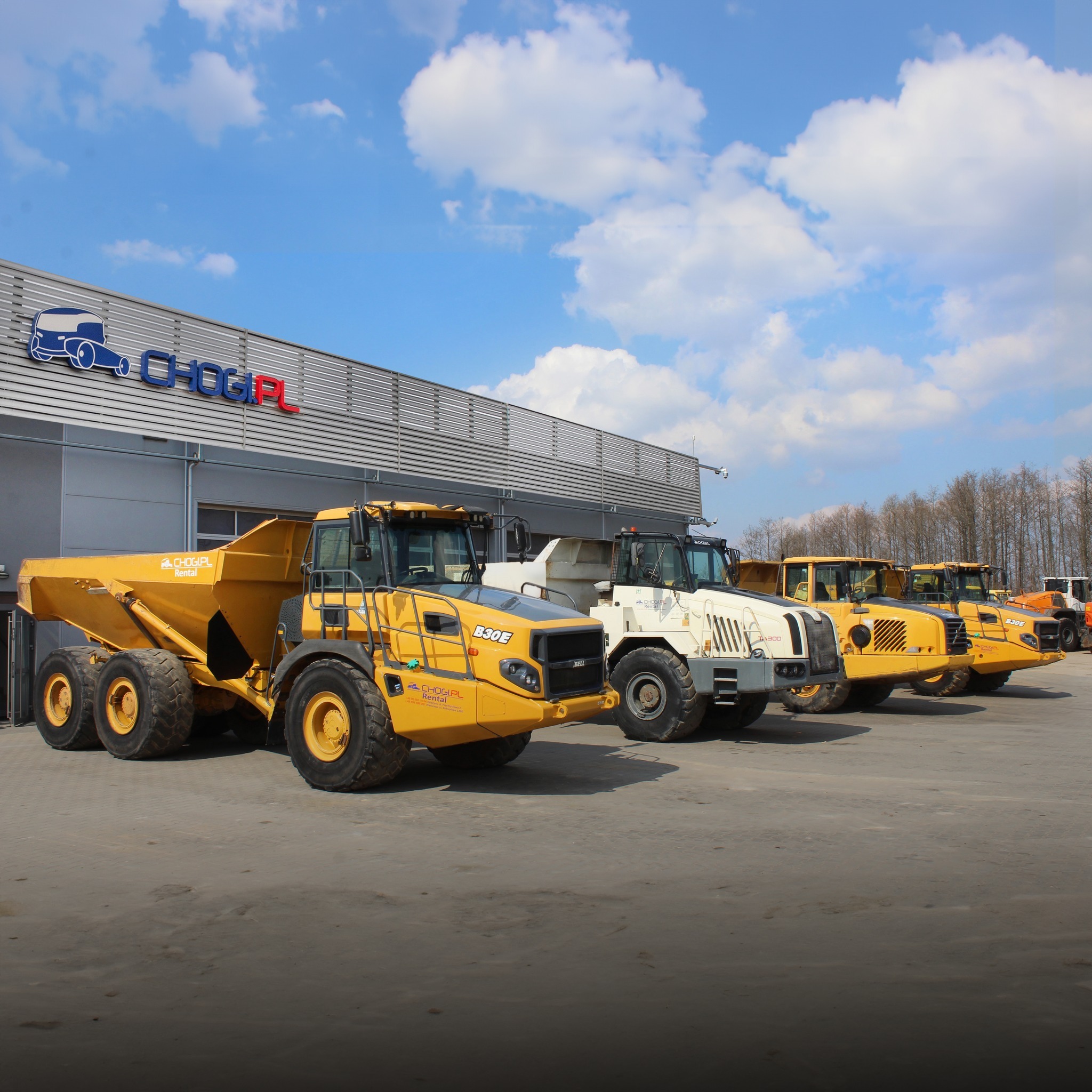Rząd żółtych i białych wywrotek przegubowych zaparkowanych przed budynkiem z logo firmy CHOGI.PL na tle błękitnego nieba z chmurami.