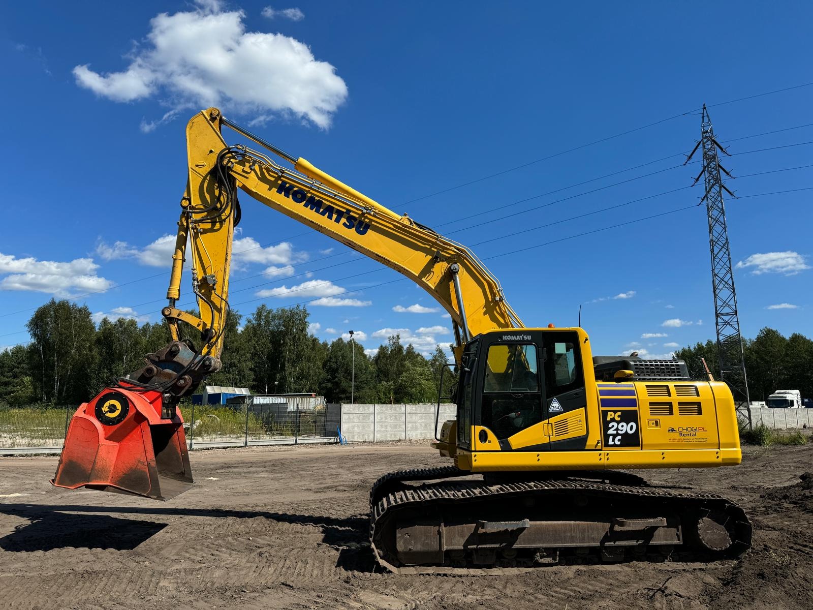 Żółta koparka Komatsu PC 290 LC z czerwonym osprzętem na tle błękitnego nieba z chmurami i słupem wysokiego napięcia.