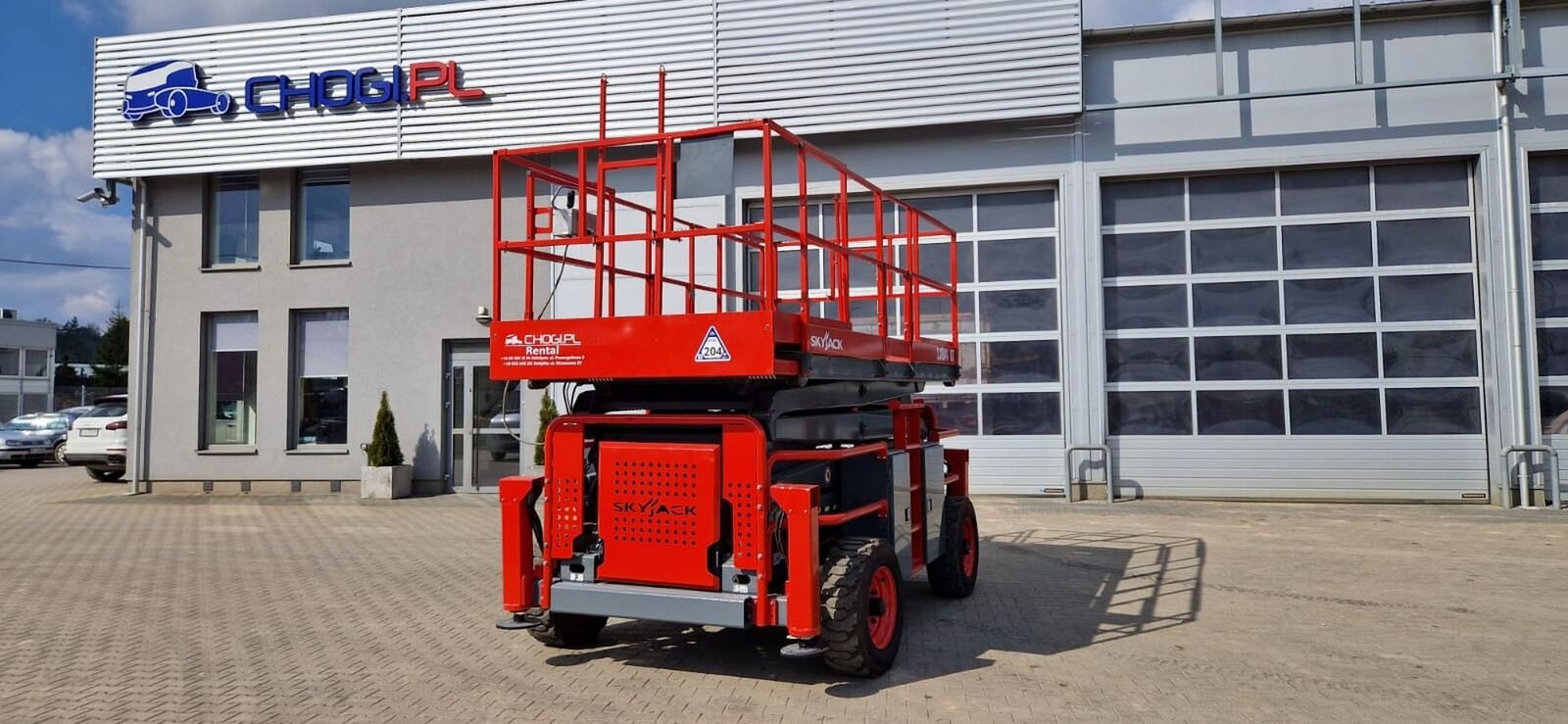 Czerwony podnośnik nożycowy SKYJACK na tle budynku z logo CHOGIPL Rental, widok z tyłu, kostka brukowa.