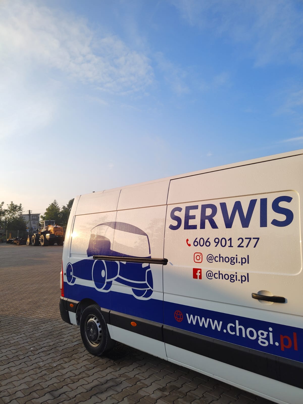Biały samochód serwisowy z niebieskim logo firmy i danymi kontaktowymi na tle placu budowy z koparką w oddali, oświetlony porannym słońcem.
