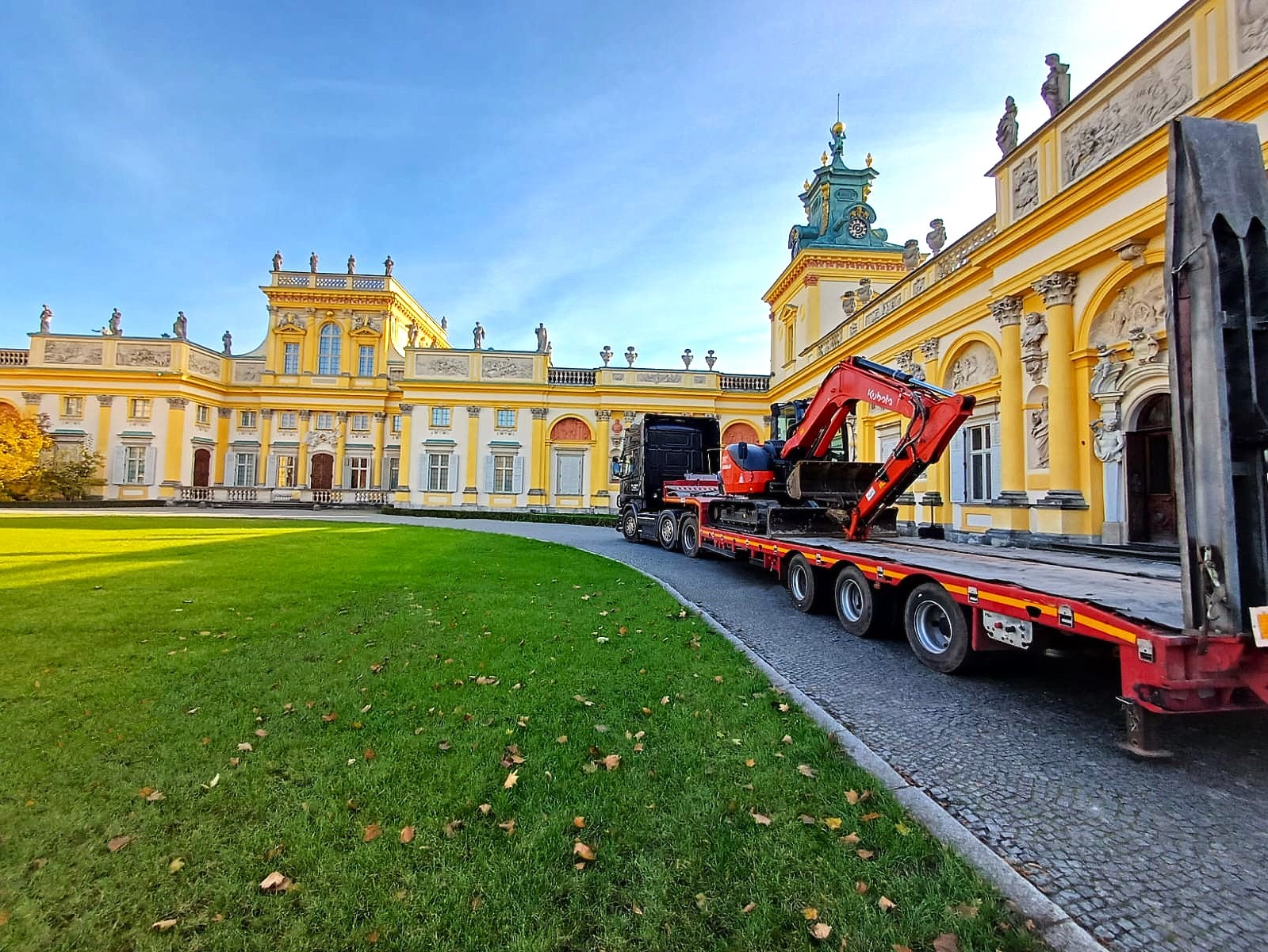 Ciężarówka z czerwoną koparką Kubota na lawecie przed Pałacem w Wilanowie, błękitne niebo
