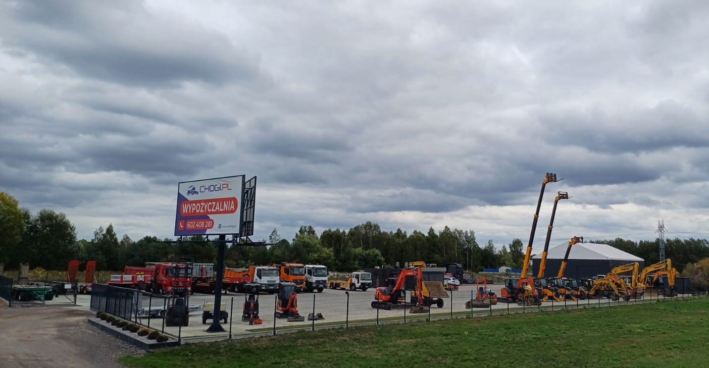 Plac wypożyczalni sprzętu budowlanego Chogi.pl z widocznymi koparkami, podnośnikami koszowymi i samochodami ciężarowymi na tle pochmurnego nieba.