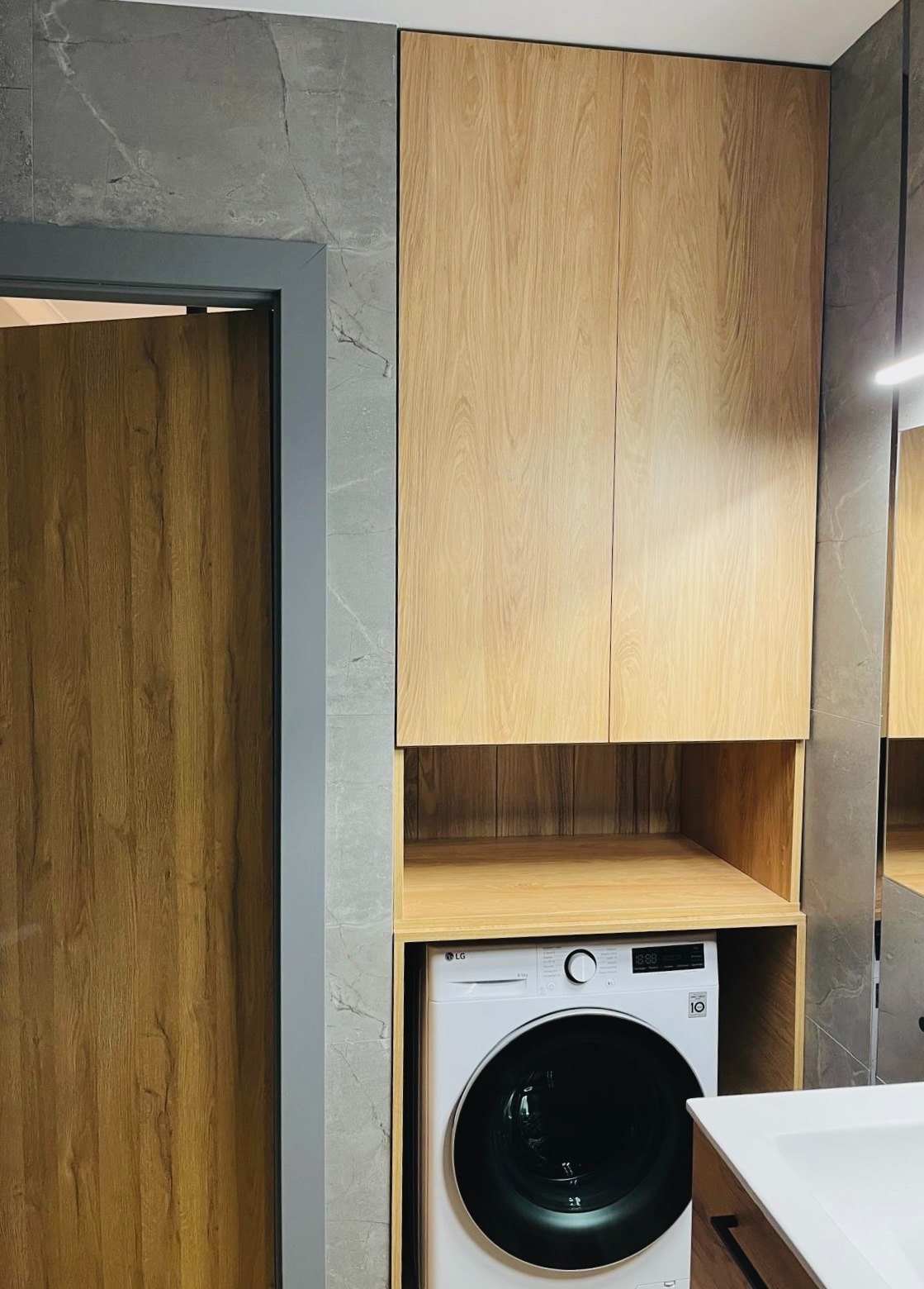 Jasne drewniane szafy na wymiar w łazience z szarą marmurową ścianą, zabudowa pralki LG.