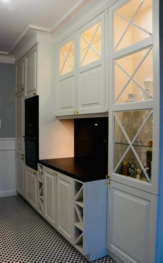 Biała zabudowa kuchenna z przeszklonymi szafkami górnymi z krzyżowymi szprosami, czarnym blatem i wbudowanym piekarnikiem, uzupełniona o otwarty regał na wino przy podłodze z czarno-białą szachownicą.