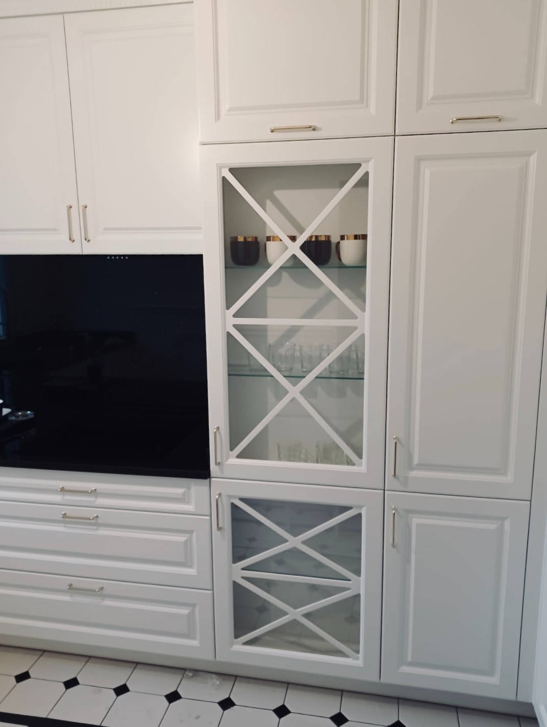 Biała zabudowa kuchenna z frezowanymi frontami, szklaną witryną z dekoracyjnymi krzyżulcami, czarnym panelem szklanym nad blatem i szufladami, na podłodze biało-czarna szachownica.