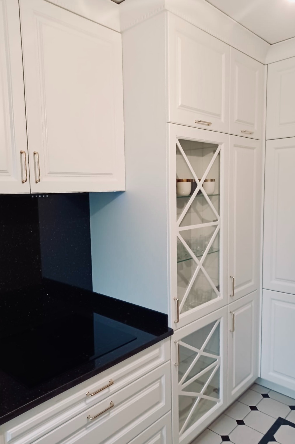 Biała zabudowa kuchenna z czarnym, błyszczącym blatem i przeszkloną witryną z dekoracyjnymi krzyżami, na podłodze płytki w czarno-białą szachownicę.