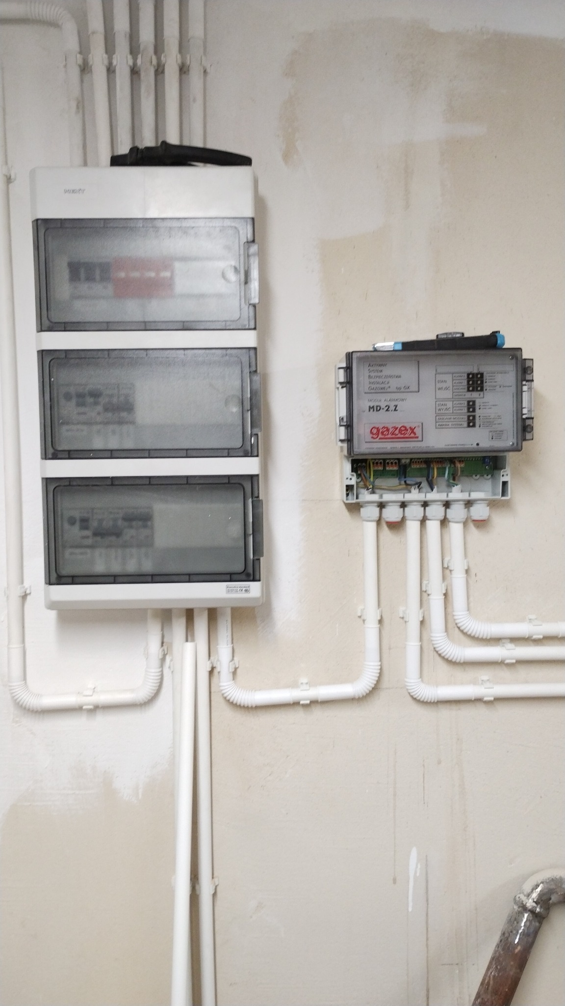 Rozdzielnica elektryczna trójsegmentowa z zabezpieczeniami i skrzynka sterownicza Gazex MD-2.2 zamontowane na ścianie z instalacją elektryczną w białych rurkach PCV.