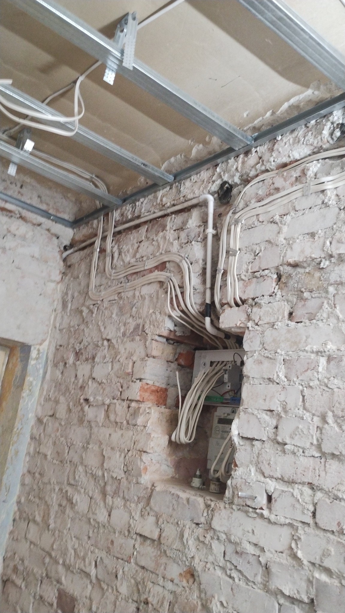 Białe przewody prowadzone po powierzchni ściany z cegły, widoczna skrzynka elektryczna wnęce, metalowa konstrukcja sufitu podwieszanego w tle.