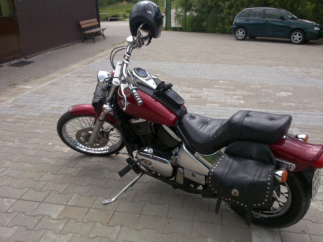 Czerwony motocykl Kawasaki VN 800 A z czarnymi skórzanymi sakwami i kaskiem na kierownicy, zaparkowany na brukowanym placu przed budynkiem, z zielonym samochodem i ławką w tle.
