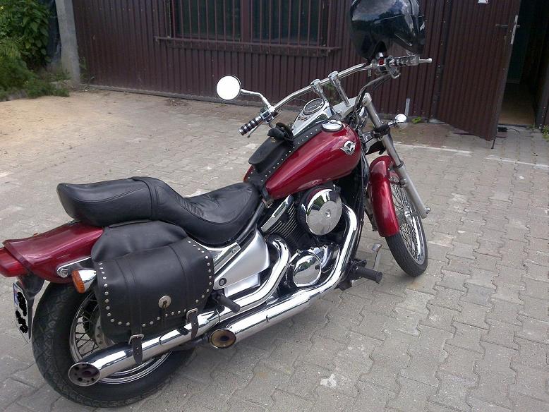 Czerwony motocykl Kawasaki VN 800 A z czarnymi sakwami, zaparkowany na szarym chodniku przed brązową bramą garażową i czarnym kaskiem na kierownicy.