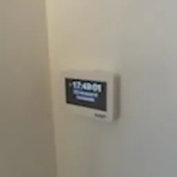 Biały panel sterowania systemu alarmowego z wyświetlaczem LCD, zamontowany na jasnej ścianie, z widocznym wskazaniem godziny i komunikatem o uzbrojeniu systemu.