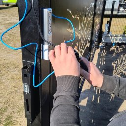 INSTALL EXPERT SPÓŁKA Z OGRANICZONĄ ODPOWIEDZIALNOŚCIĄ - Montaż domofonu przy bramie wjazdowej: elektryk podłącza przewody do panelu z klawiaturą numeryczną na czarnej, metalowej bramie z ażurowym wzorem.