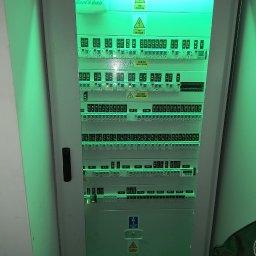 INSTALL EXPERT SPÓŁKA Z OGRANICZONĄ ODPOWIEDZIALNOŚCIĄ - Rozdzielnica elektryczna Rain Bird w obudowie, z licznymi bezpiecznikami i oznaczeniami, oświetlona zielonym światłem, z widocznymi ostrzeżeniami o wysokim napięciu.
