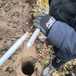 INSTALL EXPERT SPÓŁKA Z OGRANICZONĄ ODPOWIEDZIALNOŚCIĄ - Wykopany dół w ziemi, obok leżą dwie srebrne rurki, prawdopodobnie do instalacji odgromowej. Widoczne jesienne liście i fragment ubrania osoby wykonującej pracę.