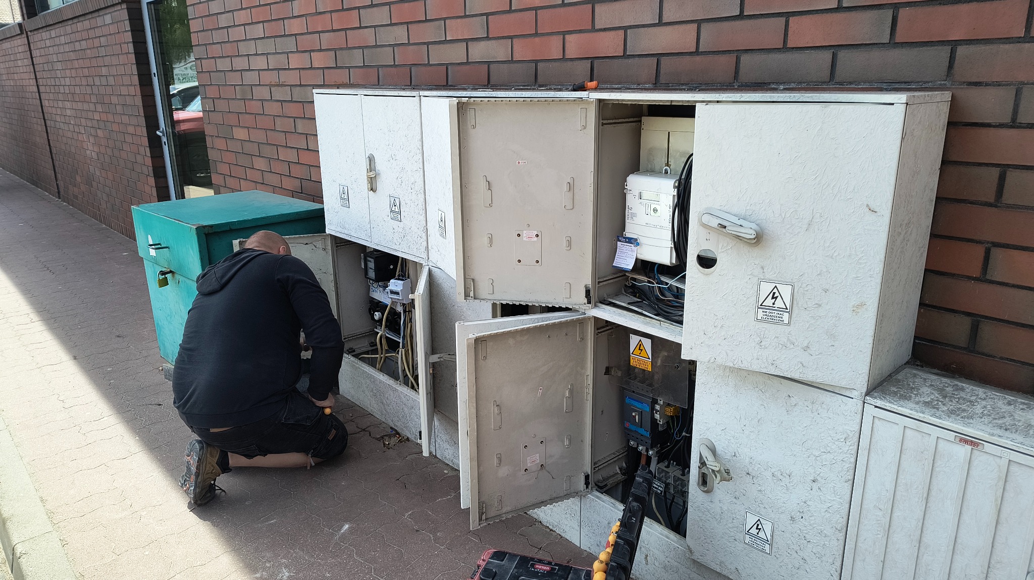 Elektryk w czarnej bluzie klęczy przed otwartą, zewnętrzną szafką elektryczną z widocznymi przewodami i urządzeniami, obok ceglany budynek.