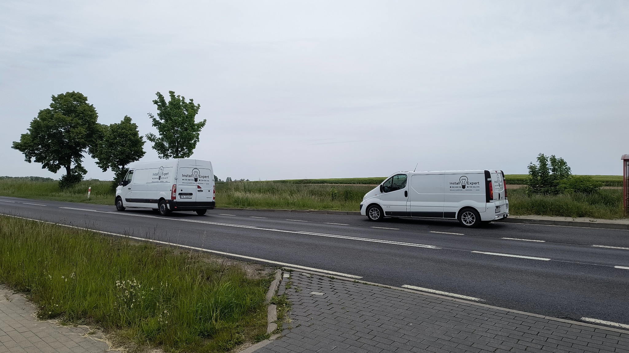 Dwa białe busy firmy Install Expert na drodze, z logo żarówki. Jeden z tyłu, drugi z boku. Trawa i drzewa w tle. Pochmurne niebo.