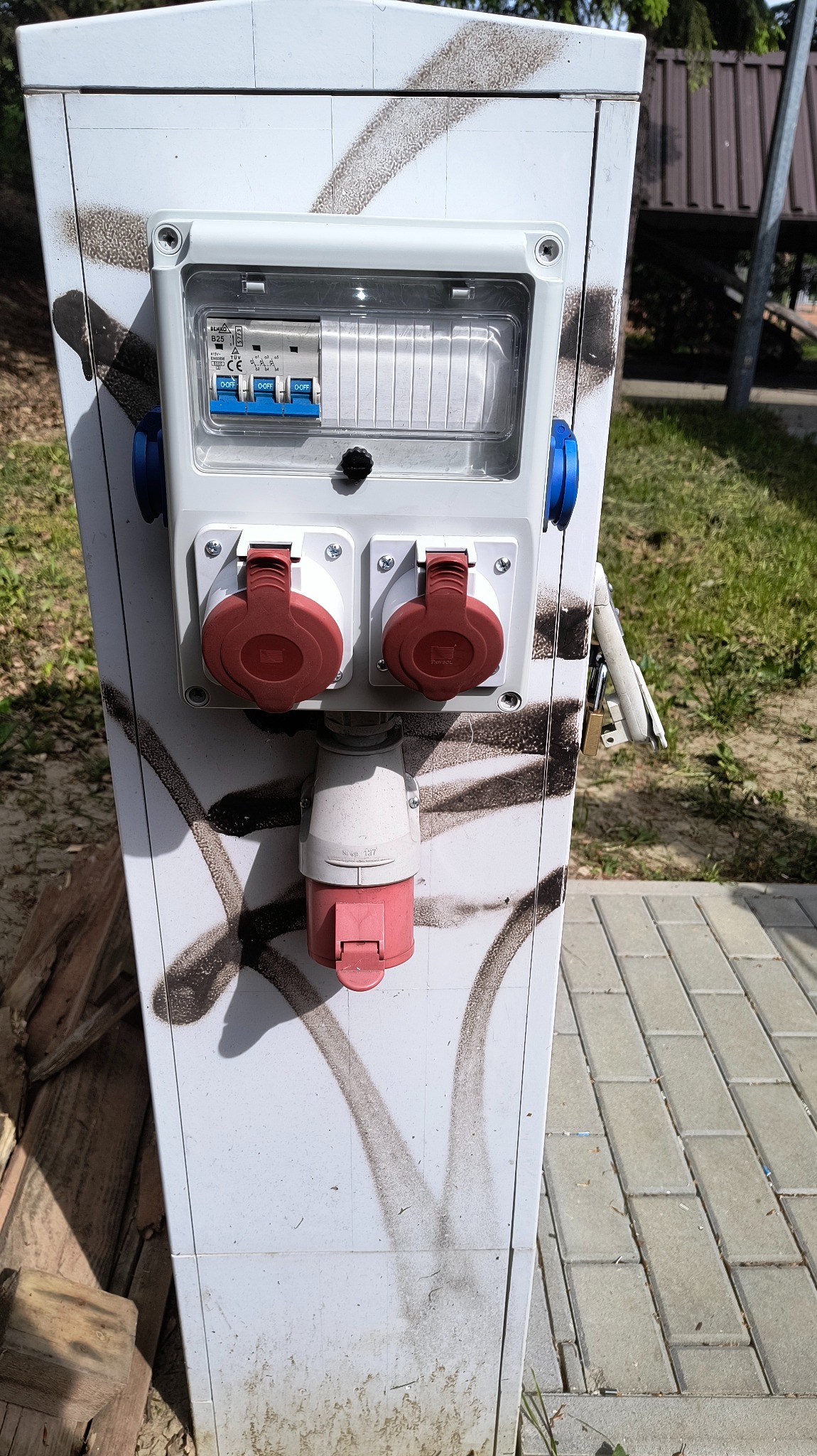 Biała skrzynka elektryczna z gniazdami siłowymi i zabezpieczeniami, umieszczona na zewnątrz, z widocznymi śladami po graffiti. Podłączony przewód z czerwoną wtyczką.