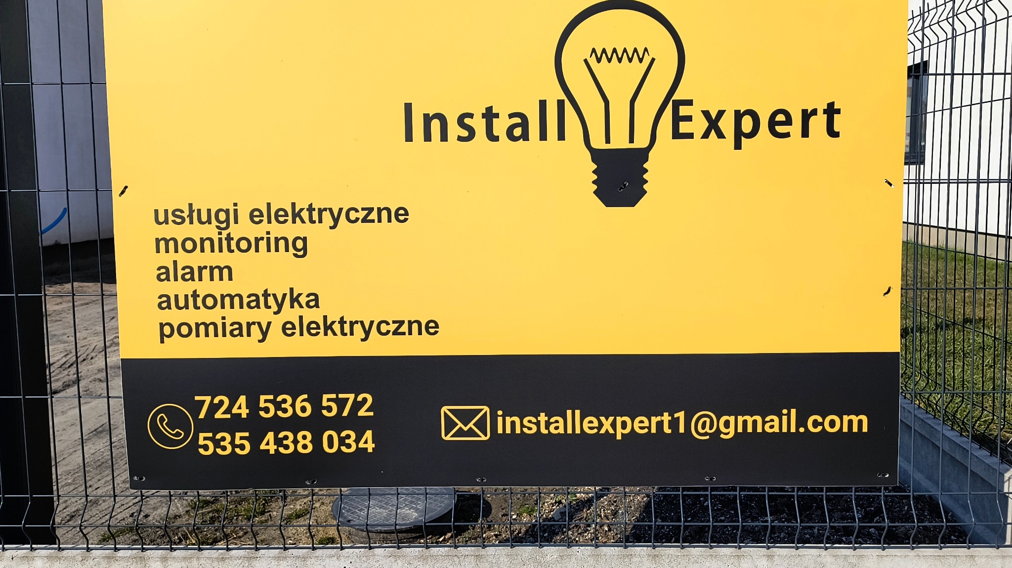 Żółto-czarny baner reklamowy firmy Install Expert z ikoną żarówki, oferującej usługi elektryczne, monitoring, alarmy i automatykę. Kontakt telefoniczny i mailowy.