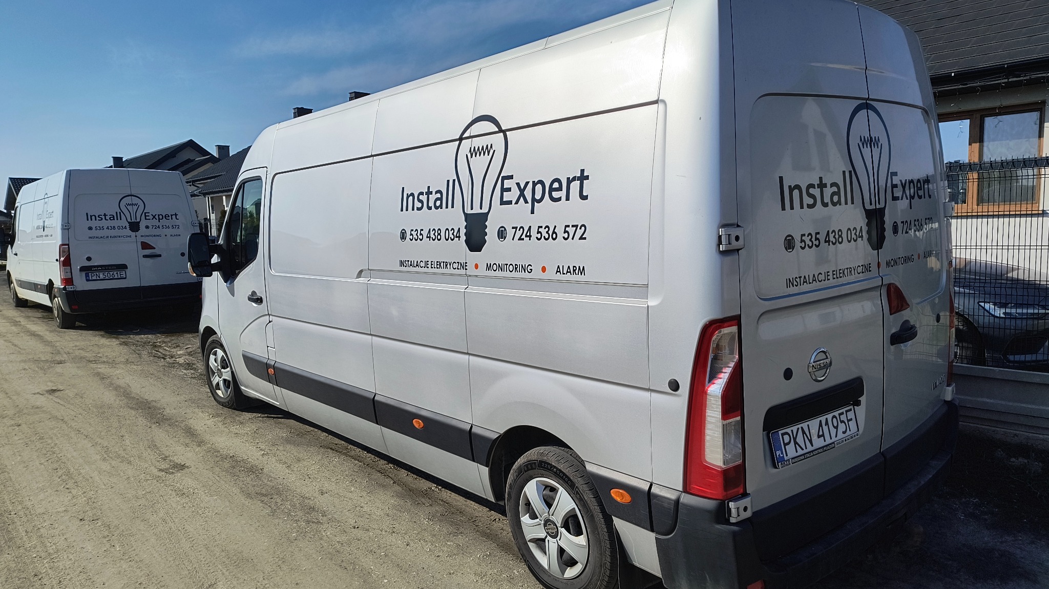 Dwa białe busy firmy Install Expert z logo żarówki, numerami telefonów i informacją o usługach: instalacje elektryczne, monitoring, alarm. Jeden bus z tablicą PKN 4195F.