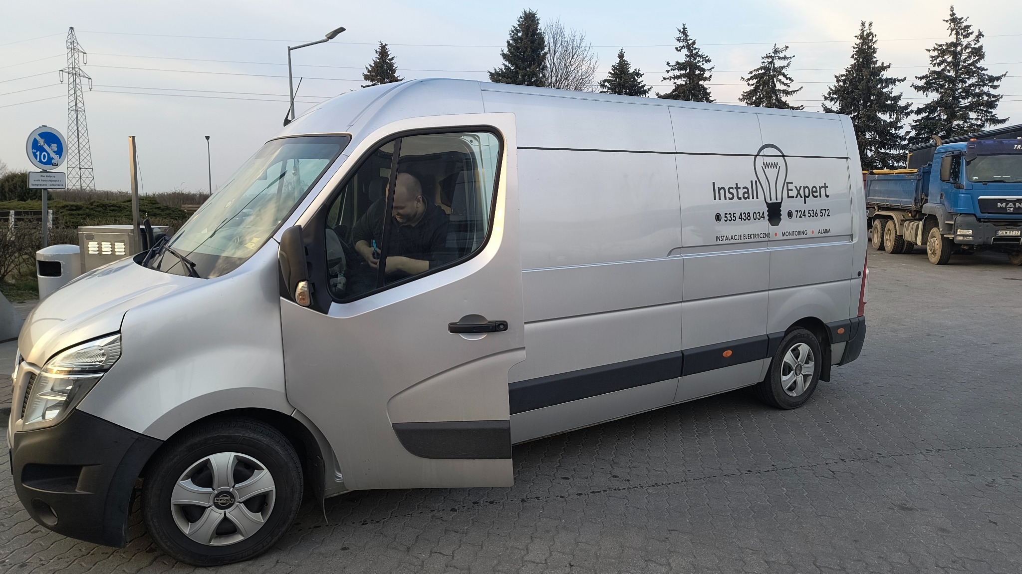 Srebrny van firmy Install Expert z otwartymi drzwiami kierowcy, zaparkowany na kostce brukowej. Na boku logo i dane kontaktowe: instalacje, monitoring, alarm.