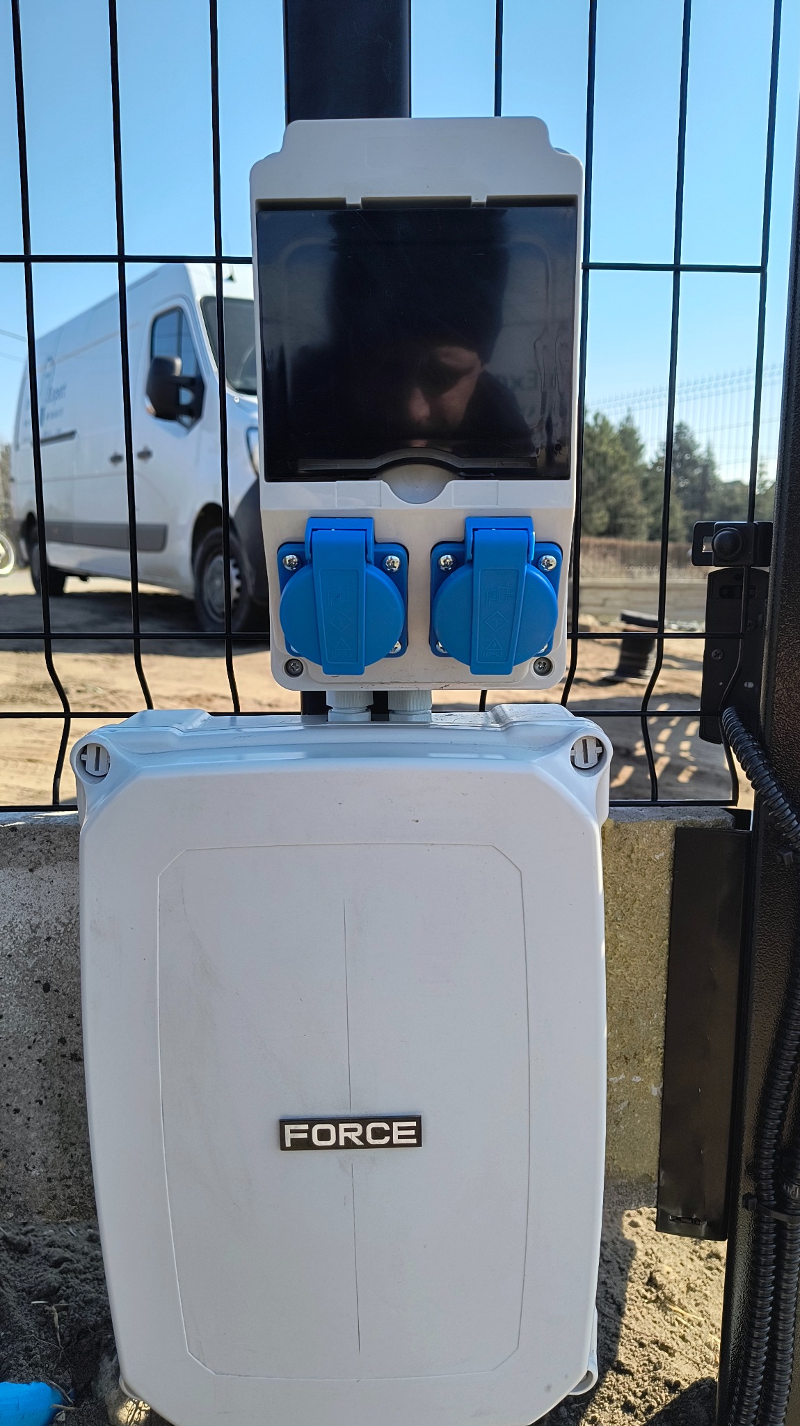 Biała skrzynka elektryczna FORCE z dwoma niebieskimi gniazdami, zamontowana na ogrodzeniu. W tle widoczny biały samochód dostawczy i fragment zieleni.