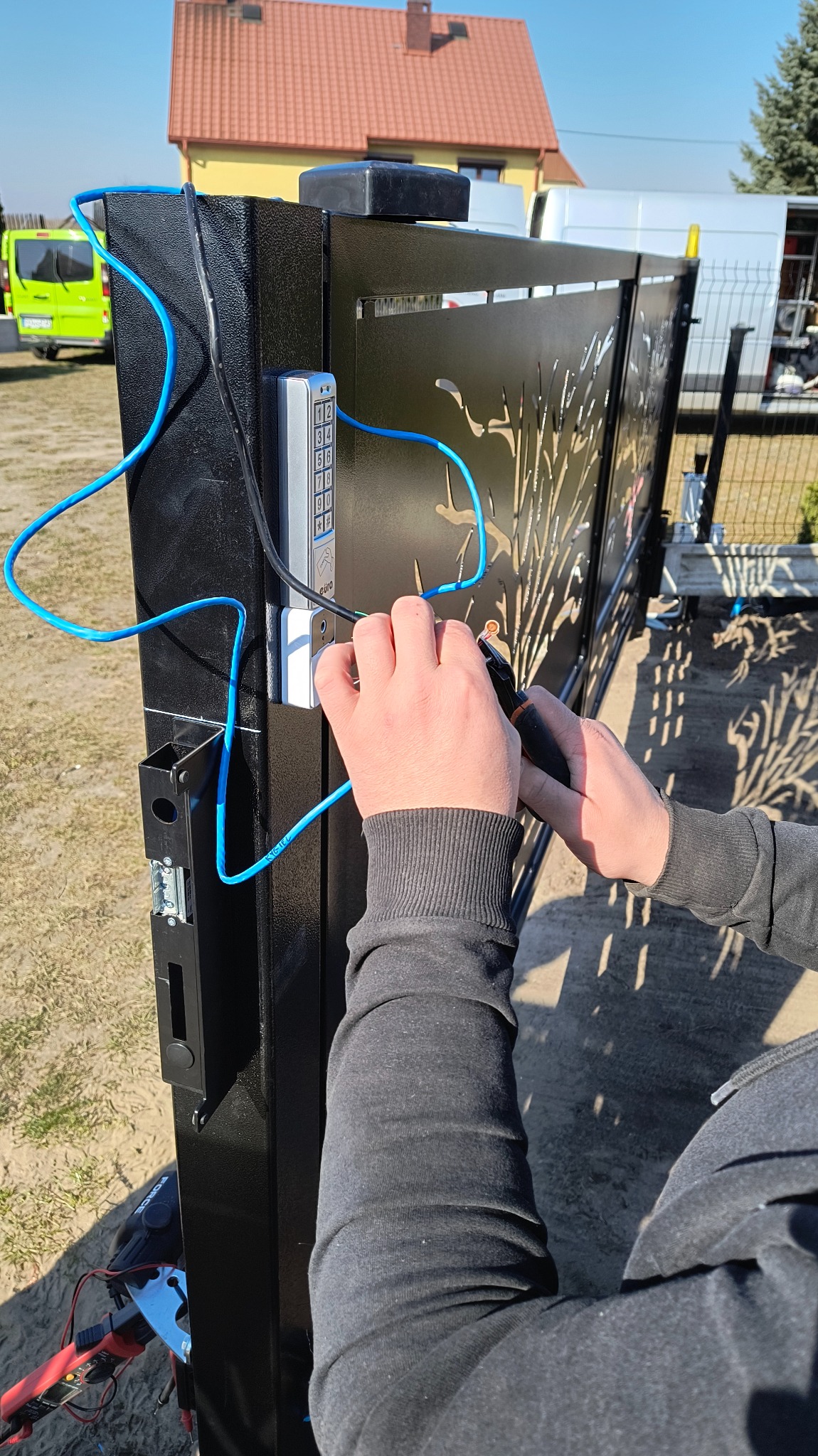 Montaż domofonu przy bramie wjazdowej: elektryk podłącza przewody do panelu z klawiaturą numeryczną na czarnej, metalowej bramie z ażurowym wzorem.