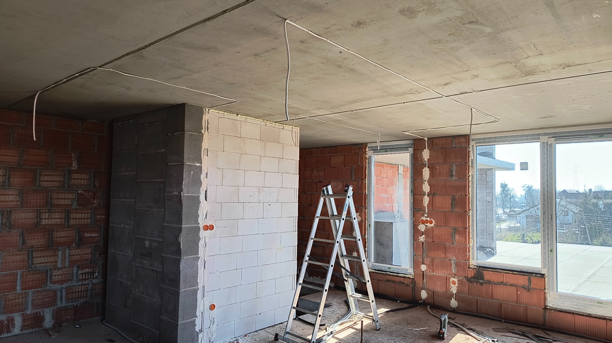 Wnętrze w stanie surowym: białe ściany z instalacją elektryczną, widoczne przewody pod sufitem, drabina. Okna z widokiem na zewnątrz. Pomieszczenie przygotowane do dalszych prac wykończeniowych.