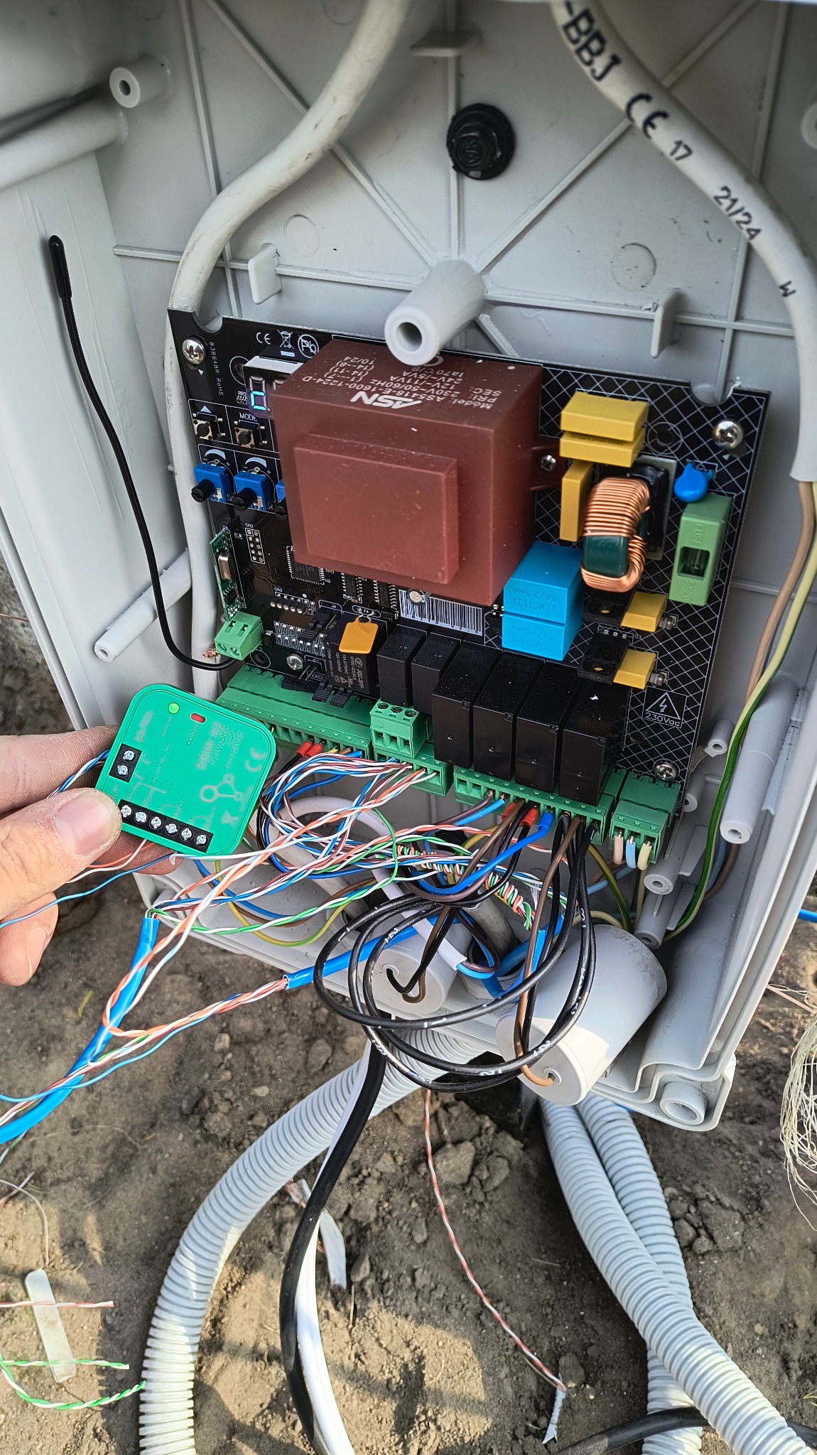 Otwarta skrzynka z płytką sterującą i licznymi przewodami, w trakcie podłączania zielonego modułu. Widoczne elementy elektroniczne i kable w peszlach.