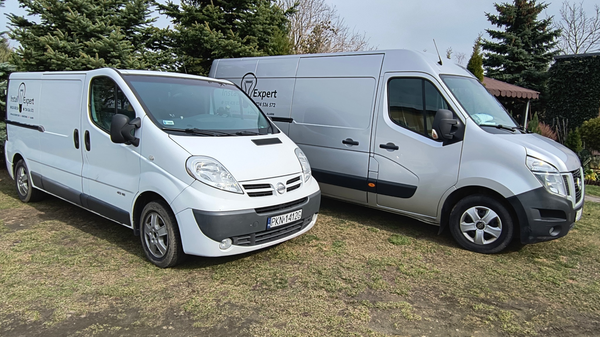 Dwa busy firmy z logo żarówki, oferującej usługi elektryczne i alarmowe, zaparkowane na trawie. Biały Nissan i srebrny Renault Master.