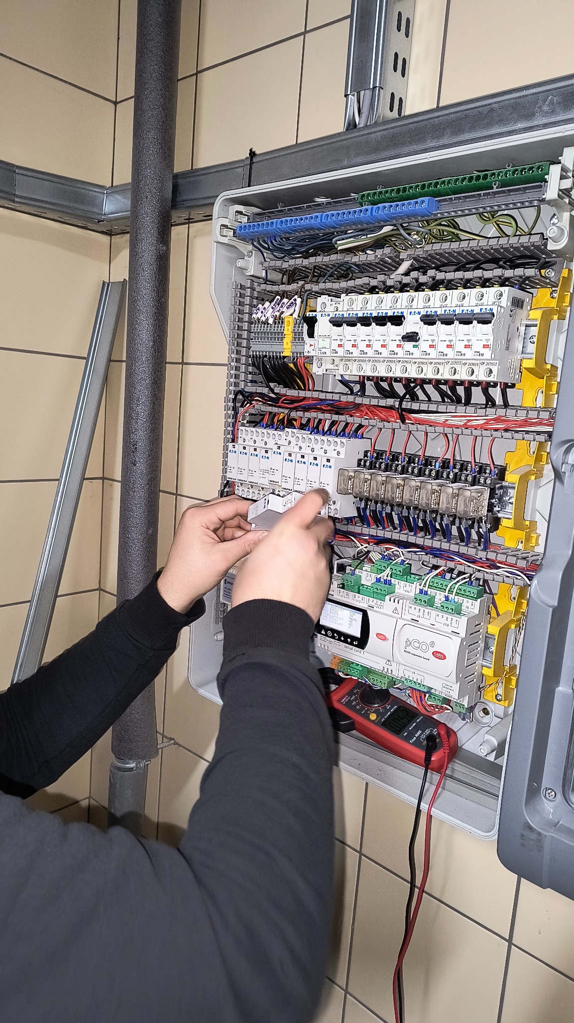 Praca przy rozdzielnicy: dłonie instalatora w czarnej bluzie montują element w skrzynce z bezpiecznikami, widoczny multimetr. Instalacja w pomieszczeniu z beżowymi płytkami.