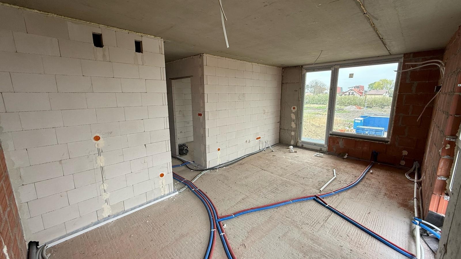 Surowy stan wnętrza z widoczną instalacją wodną (czerwono-niebieskie rury) oraz elektryczną w ścianach z betonu komórkowego i cegły, okno z widokiem na zewnątrz.