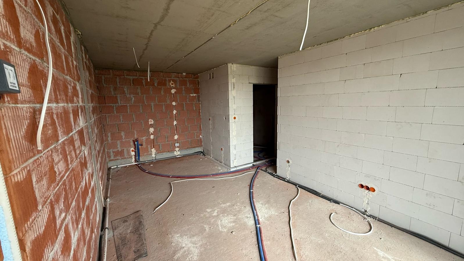Wnętrze w trakcie prac instalacyjnych: widoczne przewody elektryczne, rury wodne i ściany z cegły oraz bloczków betonu komórkowego przed wykończeniem. Stan surowy.