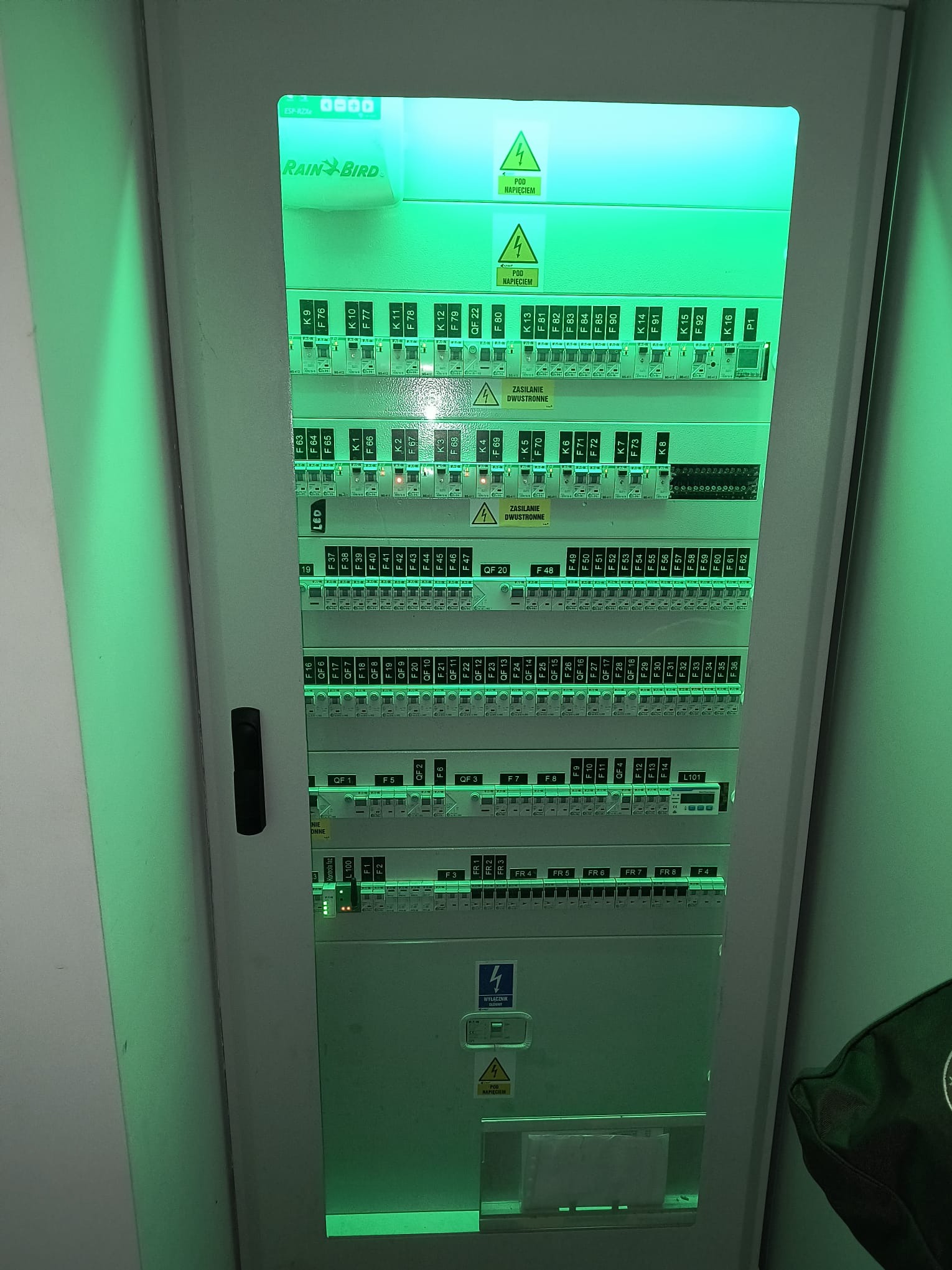 Rozdzielnica elektryczna Rain Bird w obudowie, z licznymi bezpiecznikami i oznaczeniami, oświetlona zielonym światłem, z widocznymi ostrzeżeniami o wysokim napięciu.