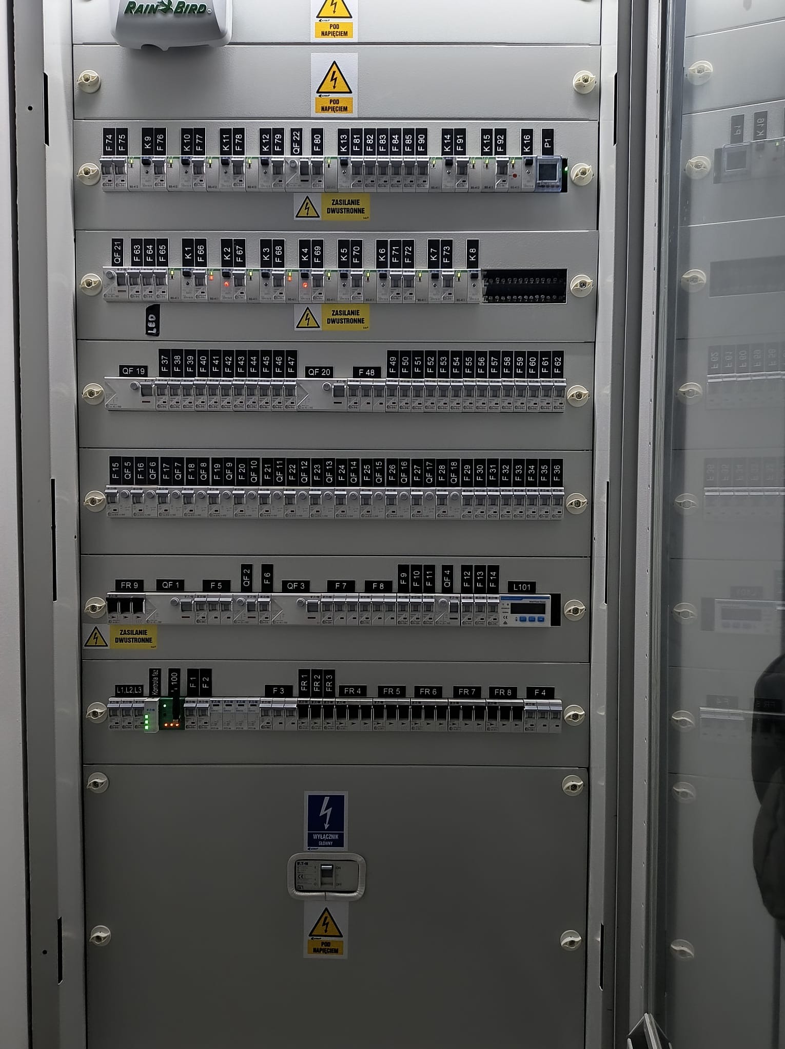Szara szafa sterownicza z bezpiecznikami, etykietami i oznaczeniami ostrzegawczymi. Widoczne liczne przełączniki i diody sygnalizacyjne. Uwagę zwraca czytelne oznakowanie.