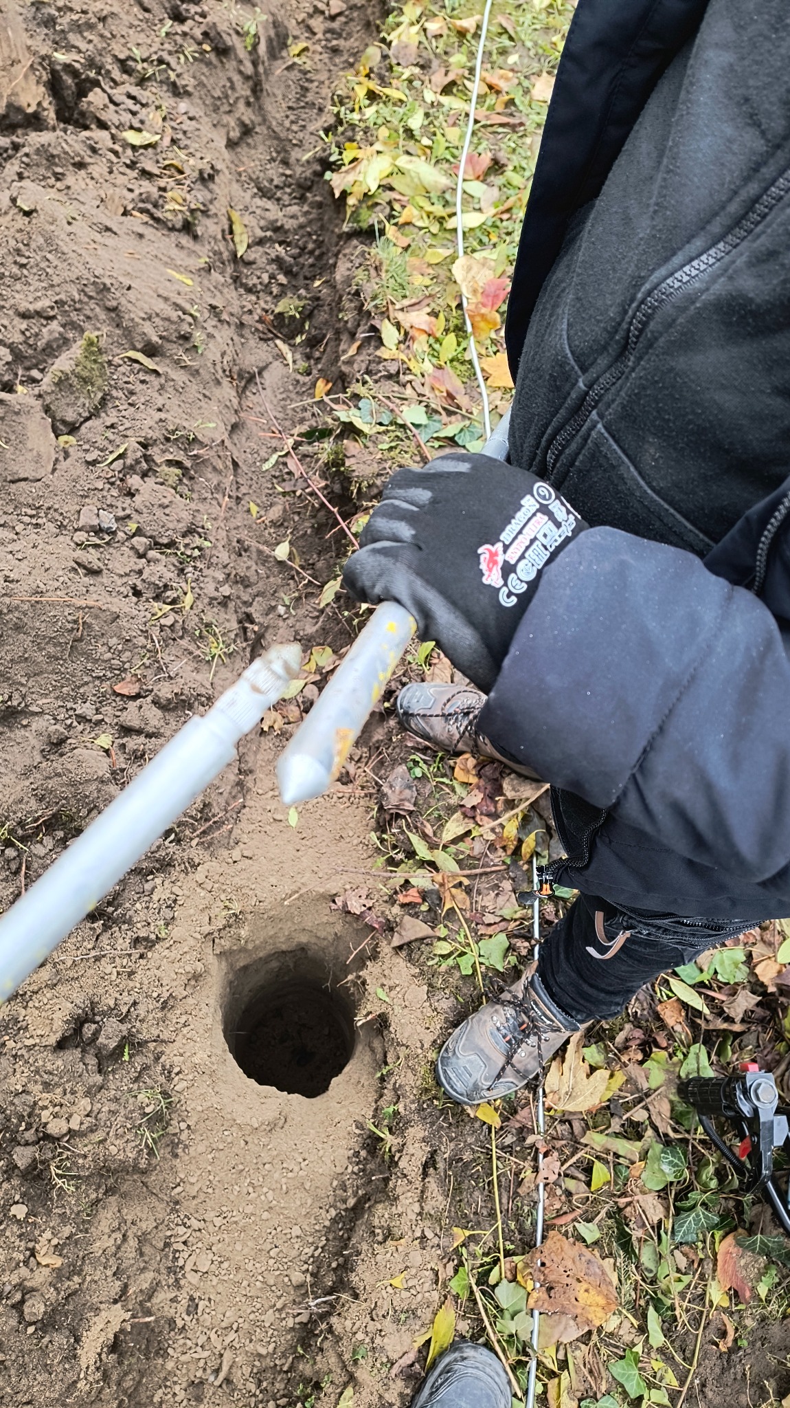Wykopany dół w ziemi, obok leżą dwie srebrne rurki, prawdopodobnie do instalacji odgromowej. Widoczne jesienne liście i fragment ubrania osoby wykonującej pracę.