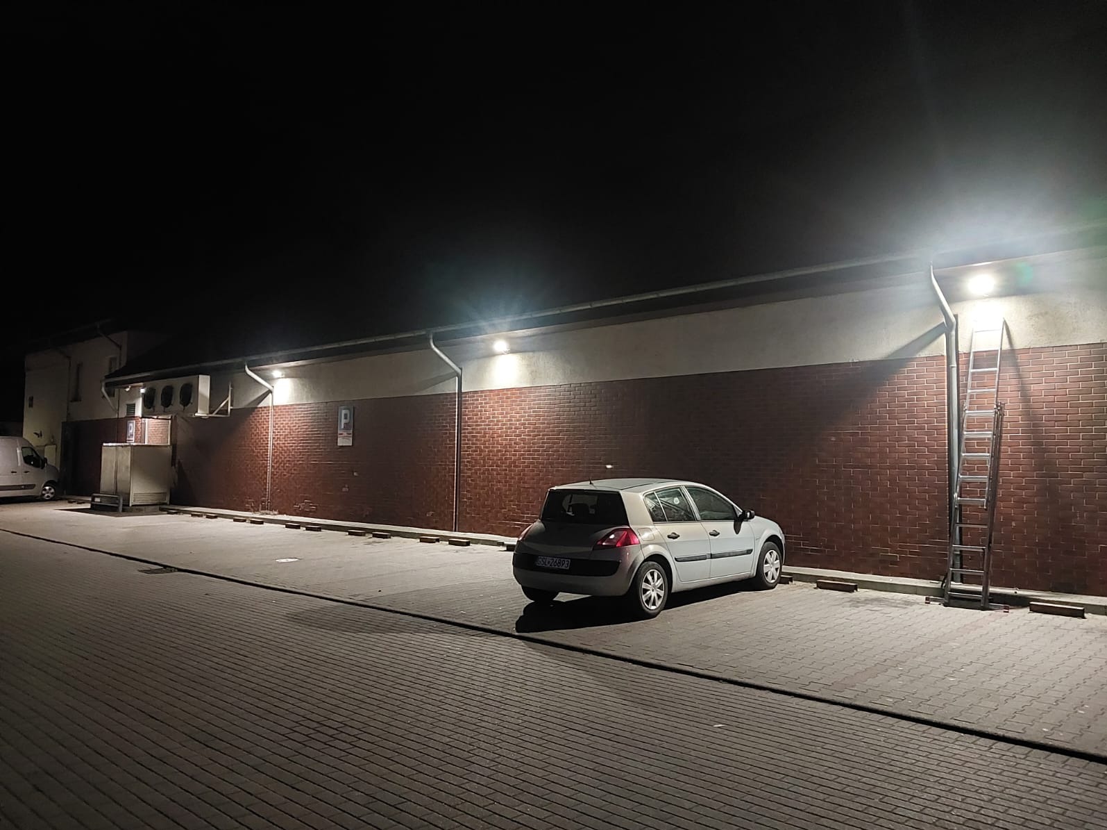 Nocny widok parkingu z szarą ścianą oświetloną lampami. Przy ścianie zaparkowany srebrny samochód i oparta drabina. Ciemna noc.