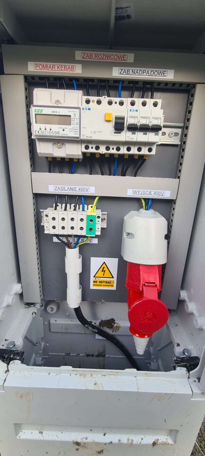 Rozdzielnica elektryczna z zabezpieczeniami, licznikiem i gniazdem 400V. Widoczne oznaczenia zasilania, wyjścia i ostrzeżenie o napięciu. Element instalacji w szarej obudowie.