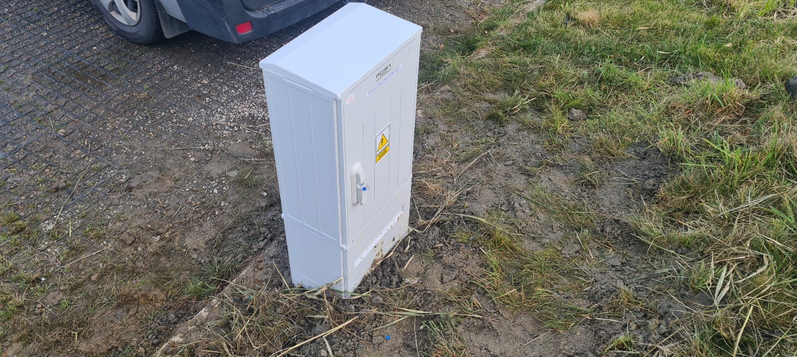 Szara szafka elektryczna z żółtym znakiem ostrzegawczym na zewnątrz, umieszczona na ziemi obok trawy i pojazdu, ukazana z perspektywy lekko z góry.