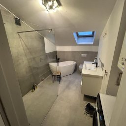 Instal Maxx - Nowoczesna łazienka na poddaszu z przeszkloną kabiną prysznicową, wolnostojącą wanną, białą umywalką i pralką, oświetlona lampą sufitową i światłem z okna dachowego.