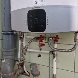 Instal Maxx - Bojler Ariston 80L z podłączonymi instalacjami hydraulicznymi w łazience z zielonymi płytkami, widoczne liczne rury z zaworami, zasilanie i odpływ.