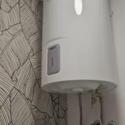 Bojler elektryczny Ariston zamontowany na ścianie z tapetą w liście, widoczne przyłącza wodne i zawory.