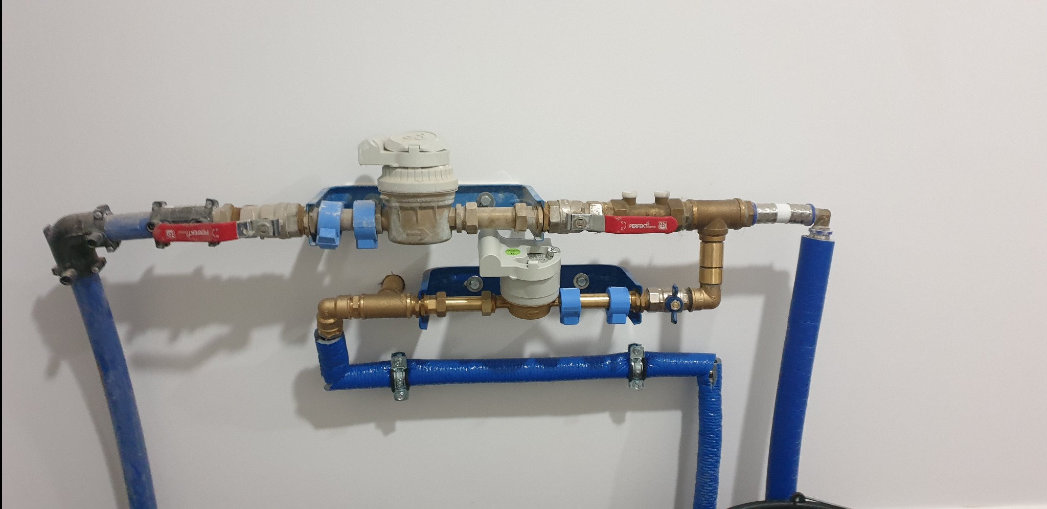 Węzeł wodomierzowy z dwoma wodomierzami na niebieskich wspornikach, z zaworami odcinającymi i niebieskimi rurami karbowanymi, zamontowany na białej ścianie.