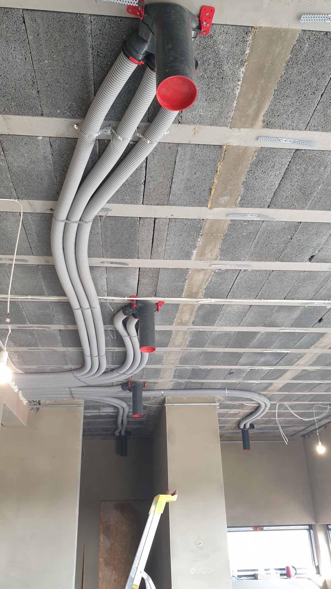 Szare, elastyczne rury wentylacyjne zamontowane pod betonowym stropem z widocznymi elementami mocującymi i czerwonymi zakończeniami, z fragmentem drabiny w dolnej części kadru.